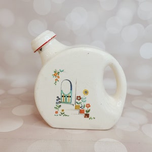 May include: White ceramic jug with a red rim and a colourful floral design featuring a blue door and yellow flowers.