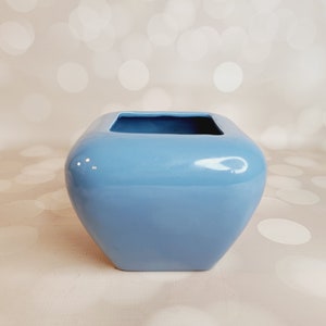 May include: A light blue ceramic planter with a square shape and rounded edges. The planter is made of glazed ceramic and has a smooth, glossy finish.