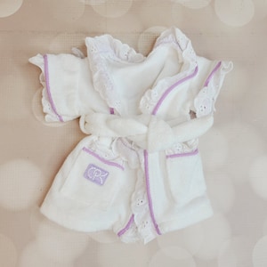 May include: White baby robe with a purple trim and lace detailing. The robe has a tie belt and a pocket with the letters "CR" embroidered on it.