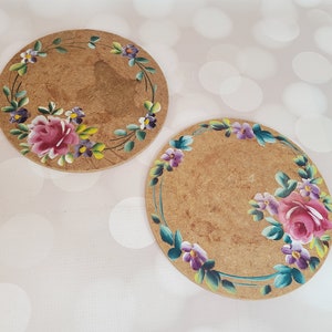 May include: Two round cork coasters with painted floral designs. The coasters feature pink, purple, and green flowers with green leaves.