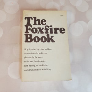 May include: A yellow book with the title "The Foxfire Book" in brown text. The book describes itself as containing information on hog dressing, log cabin building, mountain crafts and foods, planting by the signs, snake lore, hunting tales, faith healing, moonshining, and other affairs of plain living.