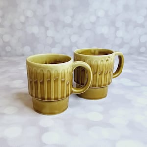 May include: Two brown ceramic mugs with a vertical ribbed design. The mugs have a rounded shape and a handle on one side.
