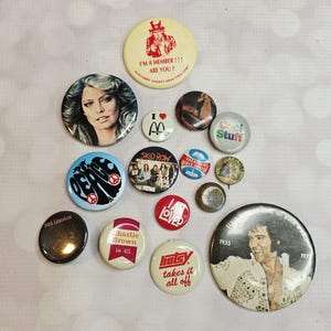 May include: A collection of vintage pinback buttons with various designs and text. The buttons feature images of celebrities, bands, and phrases like "I'm a member", "Cool Stuff", and "Loved". Colors include red, white, black, and blue.