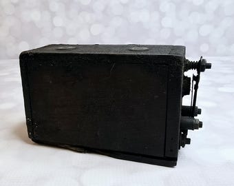 May include: A weathered, black wooden box with metal hardware and fittings. The box has a rectangular shape with visible wear and tear, suggesting age and use. The metal components include bolts, screws, and what appears to be a small lever.
