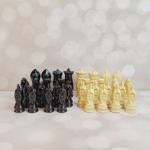 May include: A complete chess set with black and cream-colored pieces. The black pieces have a unique, stylized design, while the cream pieces feature a face motif. The set is arranged on a light-colored surface.