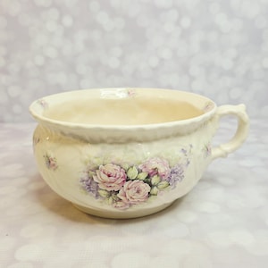 May include: A cream-colored ceramic chamber pot featuring a scalloped rim and a single side handle. The exterior is decorated with a floral motif of pink roses and small purple blossoms, with a subtle crackle glaze finish across the surface.