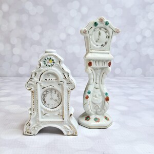 May include: Two white porcelain clock figurines. The larger clock is a tall, slender grandfather clock with a white face and black hands. The smaller clock is a more traditional style with a white face and black hands. Both clocks have painted floral details.