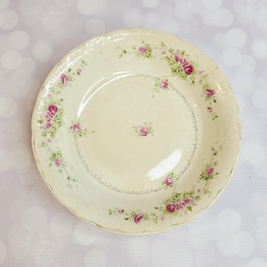 May include: Cream-colored, antique-style plate with a scalloped edge. The plate is decorated with pink rose floral patterns and green leaves. A small cluster of roses is in the center. The plate has a slightly textured surface.