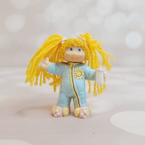 May include: A small plastic doll with yellow hair in pigtails wearing blue pajamas with a yellow sun design and holding a white bottle.