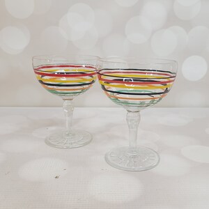 Puede incluir: Dos copas de vino de vidrio transparente con diseños de rayas coloridas. Las rayas son rojas, amarillas, verdes y negras.