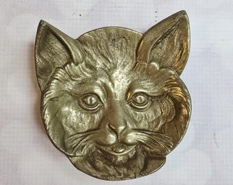 May include: A vintage metal cat face sculpture with a detailed, textured surface. The sculpture is a light gold color and features prominent ears, eyes, nose, and whiskers. The cat's expression is neutral. The sculpture is likely a decorative item.