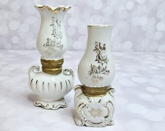 Könnte beinhalten: Zwei weiße Kerzenhalter aus Keramik mit goldenen Akzenten und floralen Designs. Die Kerzenhalter haben die Form einer Lampe mit einem breiten Fuß und einem schmalen Hals.