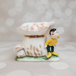 May include: A ceramic mushroom planter with a brown and white glaze. A small ceramic elf figurine with a yellow shirt and green pants is leaning against the mushroom.