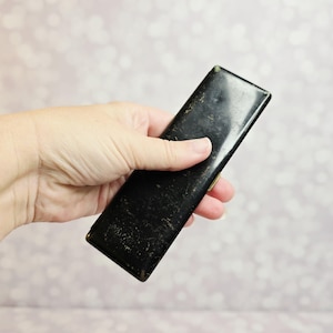 May include: A rectangular, black metal case with a smooth finish and visible signs of wear, including minor surface scratches and paint loss along the edges. The slim, elongated container features a hinged design with a metallic gold-toned clasp.
