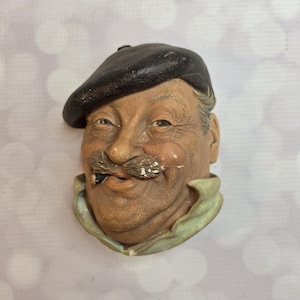 May include: A vintage ceramic wall hanging featuring a smiling man with a dark beret and a mustache. The man is wearing a light green collar. The sculpture has a weathered appearance, with visible paint wear.