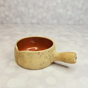 May include: A small, round ceramic bowl with a single handle extending from the side. The exterior has a textured, matte beige finish with subtle dark speckles, while the interior features a smooth, glossy, deep reddish-brown glazed surface.