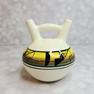 May include: A cream-coloured ceramic vase with a handle and two spouts. The vase features a geometric design in yellow, black, and brown. The vase is round with a flat base.