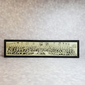 May include: A framed black and white photograph of a large group of uniformed people, likely soldiers, seated and standing in front of a building. The photograph is rectangular and has a black frame. The image is vintage and has a sepia tone.