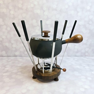 May include: A black fondue pot with a wooden handle and six metal forks on a wooden base. The pot has a silver lid and a silver burner underneath.