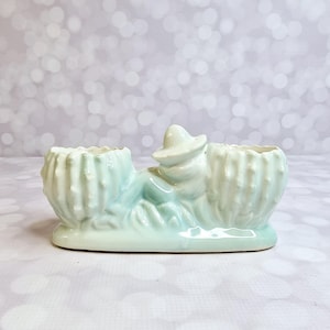 May include: A light green ceramic planter with a cowboy figure and two cactus-shaped openings. The cowboy is wearing a hat and is reclining on a bed of grass. The planter has a glossy finish.