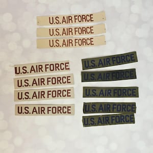 May include: A collection of rectangular fabric name tapes featuring the text U.S. AIR FORCE embroidered in brown on tan fabric and in dark blue on olive green fabric. These patches are arranged in three distinct groups on a light background.