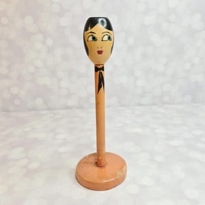 May include: A vintage wooden hat stand featuring a painted face with black hair, blue eyes, and red lips. The stand has a slender, cylindrical stem and a round base, painted in a light peach color. A black bow is painted below the face.