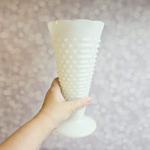 Könnte beinhalten: Eine weiße Hobnail-Vase mit ausgestellter Oberseite und rundem Sockel. Die Vase hat eine strukturierte Oberfläche mit kleinen, erhabenen Noppen. Die Vase wird von einer Hand gehalten.