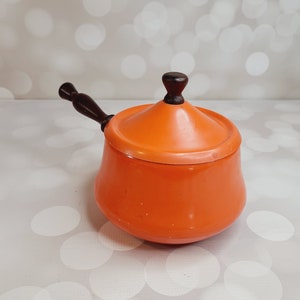 Puede incluir: Cacerola de esmalte naranja con mango de madera y tapa. La cacerola está sobre un fondo blanco.