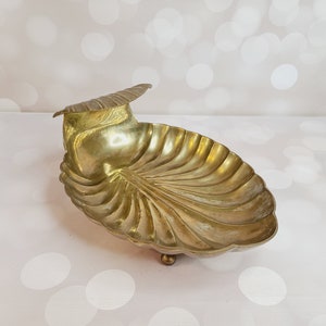 May include: A gold-toned brass shell-shaped dish with a raised lip and three feet. The dish is designed to resemble a seashell.