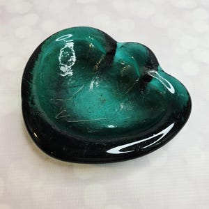 May include: A teal-colored, heart-shaped glass dish. The dish has a glossy finish and a slightly uneven surface, with a darker rim. The heart shape is clearly defined, and the glass appears to be transparent, allowing light to pass through.
