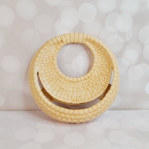 Puede incluir: Un objeto decorativo redondo, tejido, de color crema, con un gran agujero en el centro. El objeto está hecho de un material que se asemeja a la paja o la mimbre.
