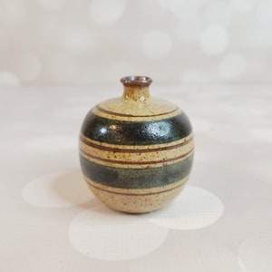 Puede incluir: Un pequeño jarrón redondo de cerámica con un patrón de rayas marrones y verdes. El jarrón tiene un esmalte moteado y una abertura ligeramente redondeada.