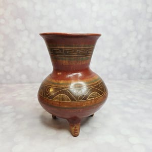 Puede incluir: Un jarrón de cerámica artesanal, de color marrón rojizo, con un cuerpo bulboso y un borde acampanado. El jarrón presenta bandas decorativas con patrones geométricos y una base de tres patas con caras talladas. El jarrón mide unos 15 cm de altura.