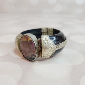 Puede incluir: Un brazalete negro y plateado con una piedra grande, ovalada y multicolor en el centro. El brazalete tiene un cierre con bisagra.