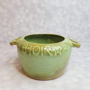 Puede incluir: Un cuenco de cerámica verde claro con dos asas pequeñas. El cuenco tiene una abertura ancha y una banda decorativa con formas y texto en relieve alrededor del borde. El exterior del cuenco tiene un degradado de verde y marrón.