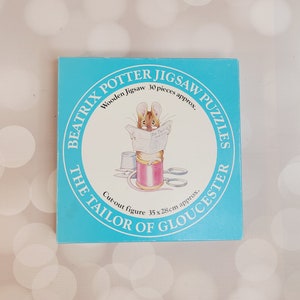 May include: A blue and white box featuring a circular image of a mouse holding a newspaper that reads "The Tailor of Gloucester". The box is for a 30-piece wooden jigsaw puzzle of Beatrix Potter's "The Tailor of Gloucester".