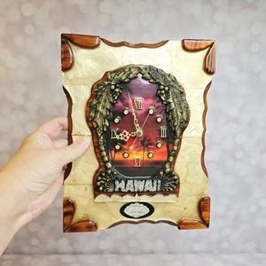 May include: A rectangular wall clock featuring a sunset scene with palm trees and the word HAWAII in raised letters. The clock face is framed by a textured cream-colored border with dark wood corners and gold-toned Roman numeral markers.