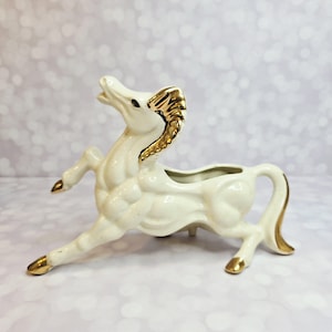 May include: A ceramic horse figurine in a running pose, featuring a cream-colored body with gold accents on the mane, hooves, and tail. The figurine has a hollowed-out area, suggesting it could be used as a planter or decorative piece.