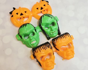 May include: A collection of Halloween-themed plastic masks. The masks include two orange jack-o'-lanterns, two green Frankenstein monster faces, and two orange Frankenstein monster faces with black hair. The masks are on a white surface.