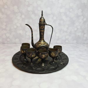 May include: An antique brass tea set with a decorative floral design. The set includes a tall, ornate teapot with a curved spout and handle, six small glasses, and a round tray. The set is in a vintage style.