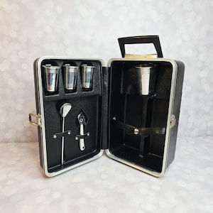 May include: A vintage black bar set in a suitcase-style case. The case is open, revealing stainless steel shot glasses, a cocktail shaker, and bar tools. The case has a black handle and silver trim.