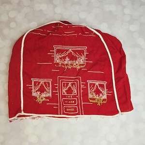 May include: A red fabric appliance cover featuring white embroidered outlines of a house with three windows, flower boxes, and a central door. The door includes the embroidered text THE MIXER HOUSE. The cover has white piping along the edges.