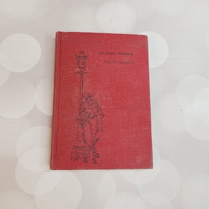 May include: A red hardcover book with the title "La Rana Viajera" and the author "Julio Camba" printed on the front cover. The cover features a black and white illustration of a street lamp with a figure standing beneath it.
