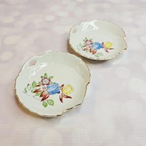 May include: Two off-white, leaf-shaped ceramic dishes with gold trim. Each dish features a floral design with blue, yellow, red, and pink flowers. The dishes have a small handle and are set against a soft pink background.