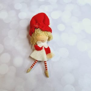 Peut inclure: Une petite poupée en feutre avec un chapeau rouge, une robe blanche et des jambes rayées rouge et blanc. La poupée a des cheveux en laine blonde et un visage peint.