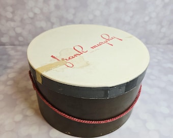 May include: A round, vintage hat box with a cream-colored lid and dark brown sides. The lid features the name "frank murphy" in red script. A red rope detail encircles the box, adding a decorative touch. The box is likely used for storing hats.
