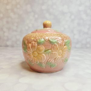 May include: A small, round, pink ceramic jar with a lid. The jar is decorated with embossed yellow and green flowers. The lid has a small, round knob. The jar has a crackle glaze finish.