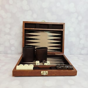 May include: Open wooden backgammon set with a brown exterior and a playing surface of alternating light and dark triangles. Includes two brown leather dice cups, dice, and game pieces. The set is displayed on a white surface.