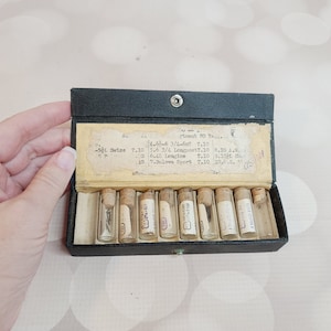 May include: A black box with a hinged lid containing small glass vials with watch parts. The box has a label that reads "Adjustment 80 Years" and lists different watch brands and sizes.