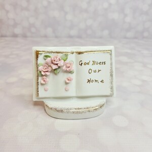 May include: A white ceramic book-shaped figurine with gold trim and the words "God Bless Our Home" in gold lettering. Pink roses and green leaves adorn the left side. The base is also white with gold accents.